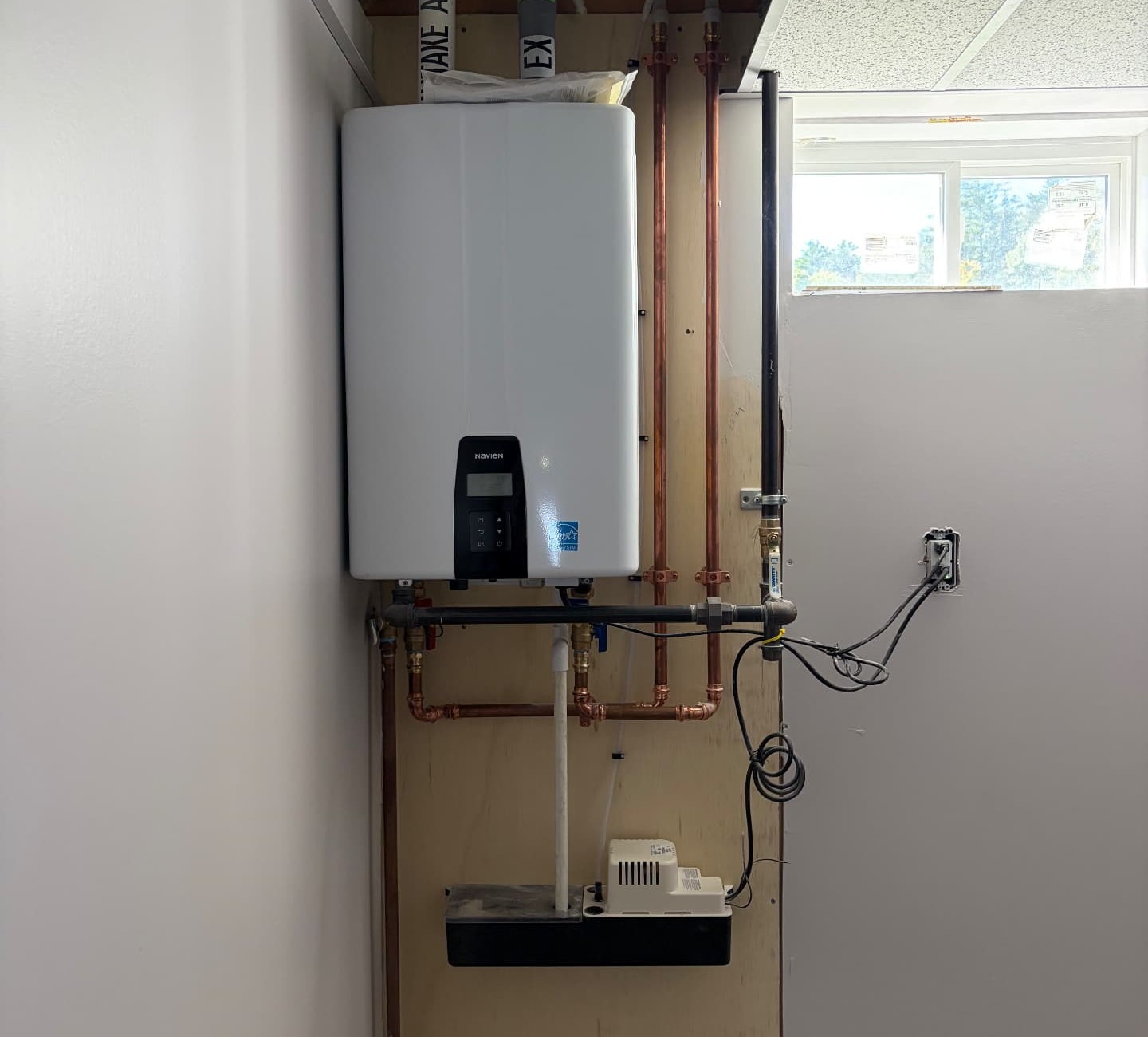 Navien tankless water heater mounted on a plywood backer with intake and exhaust venting, copper supply lines, a black iron gas line, and a condensate pump below, installed in a Charlton, MA basement