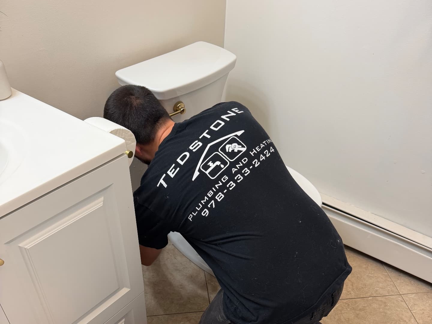 Adam Tedstone kneeling between a vanity and a toilet in a customer's bathroom, the TEDSTONE Plumbing and Heating logo and phone number readable on the back of his shirt